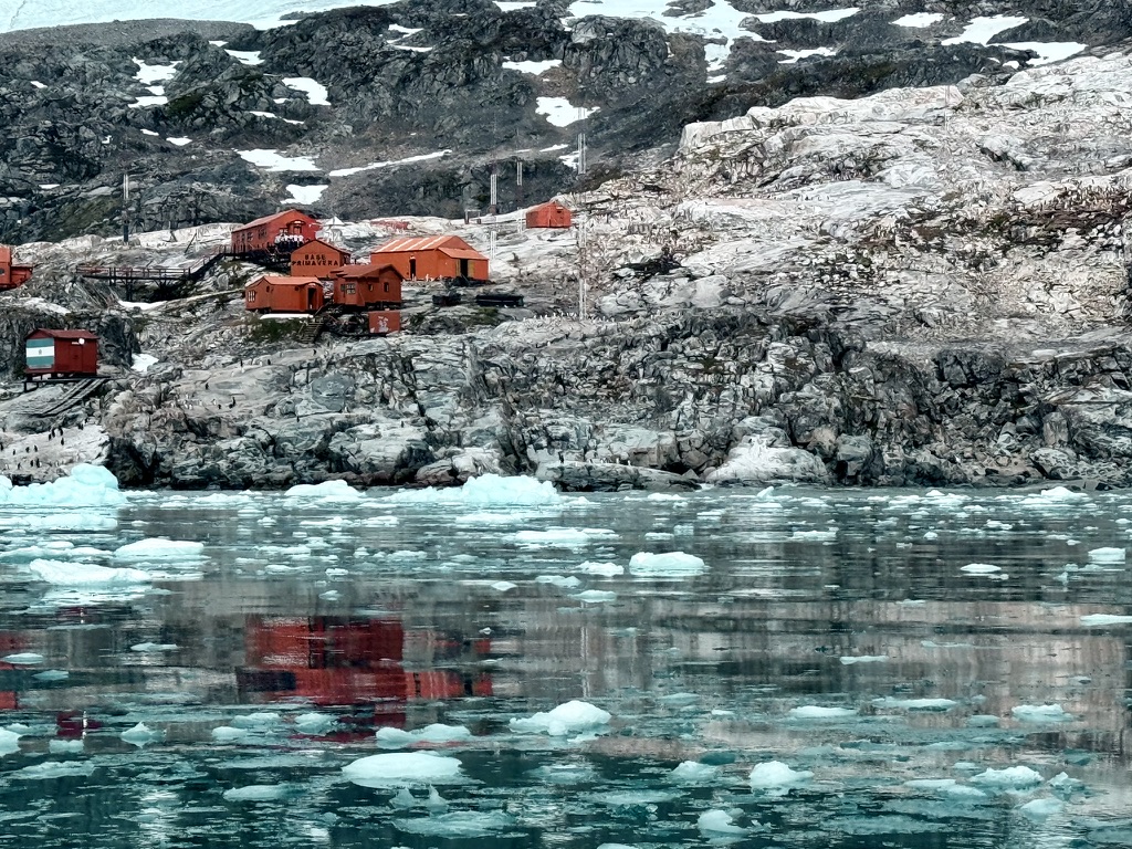 Red buildings on a rocky Antarctic shoreline reflected in water and floating ice