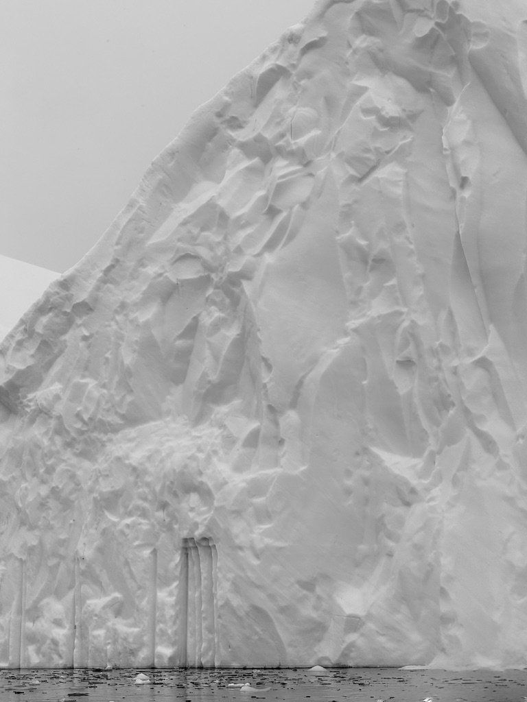 The towering white face of an Antarctic iceberg, marked by striations and deeply carved vertical forms